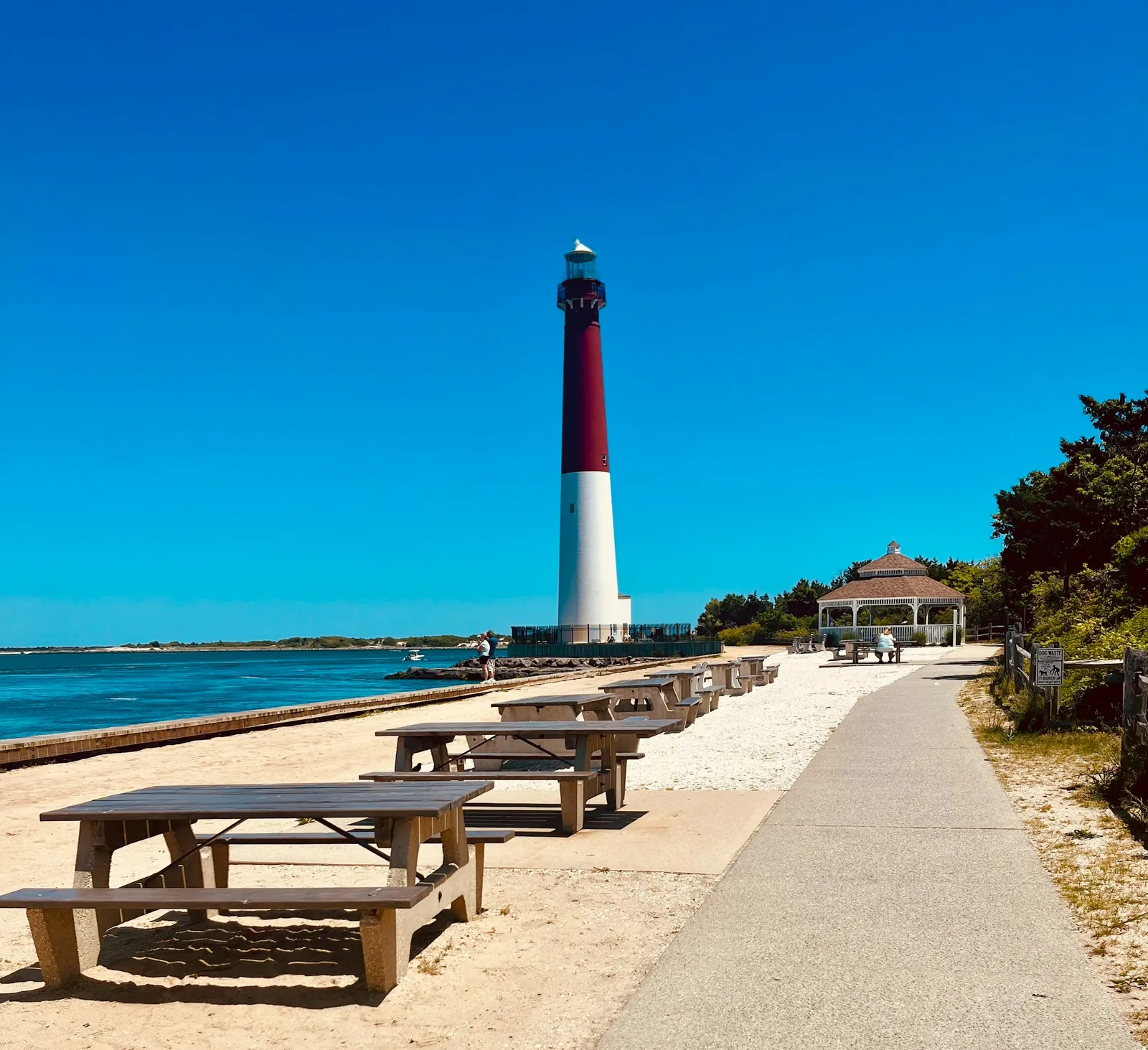 A red and white lighthouse at the end of a sunny beachside path lined with picnic tables.