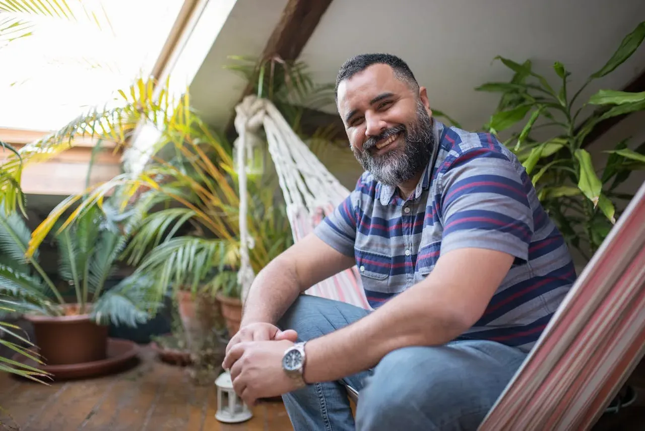 A bearded man smiling in a hammock chair in a sunlit, plant-filled sunroom.