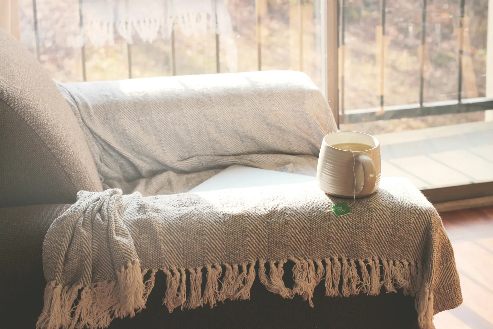 A knitted blanket and a mug on a sofa beside a bright window.
