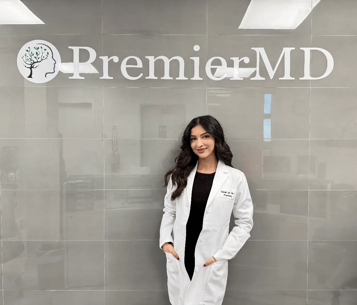 Zainab Ali, PA-C, in her white coat, smiling in front of the PremierMD sign at our office.