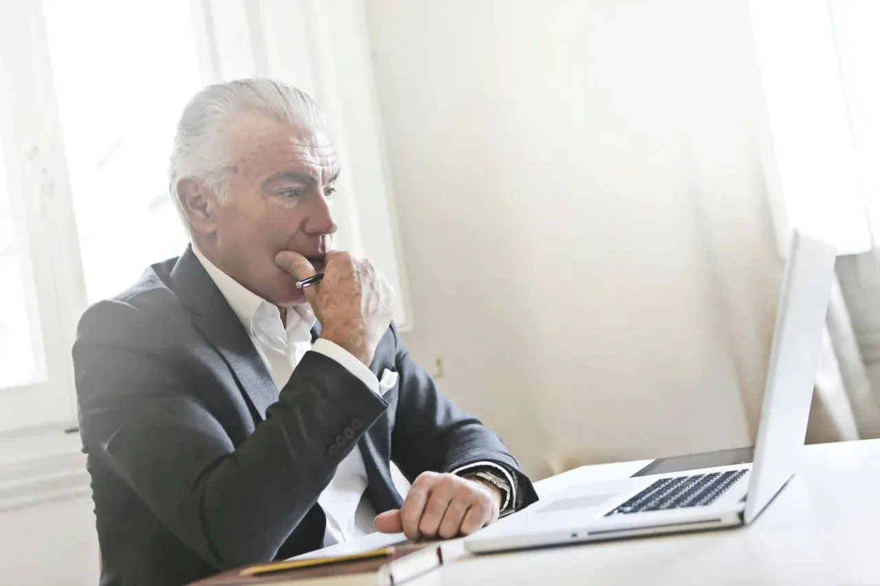 A man in a gray suit sitting at a white desk, hand to his chin, looking at an open laptop.