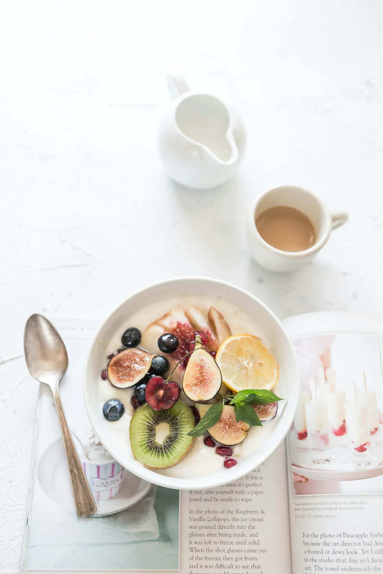 A bowl of yogurt and fruit beside a cup of coffee on a white table.