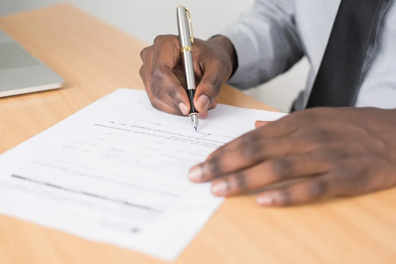 Hands in a business shirt signing a printed form with a silver pen, a laptop nearby.