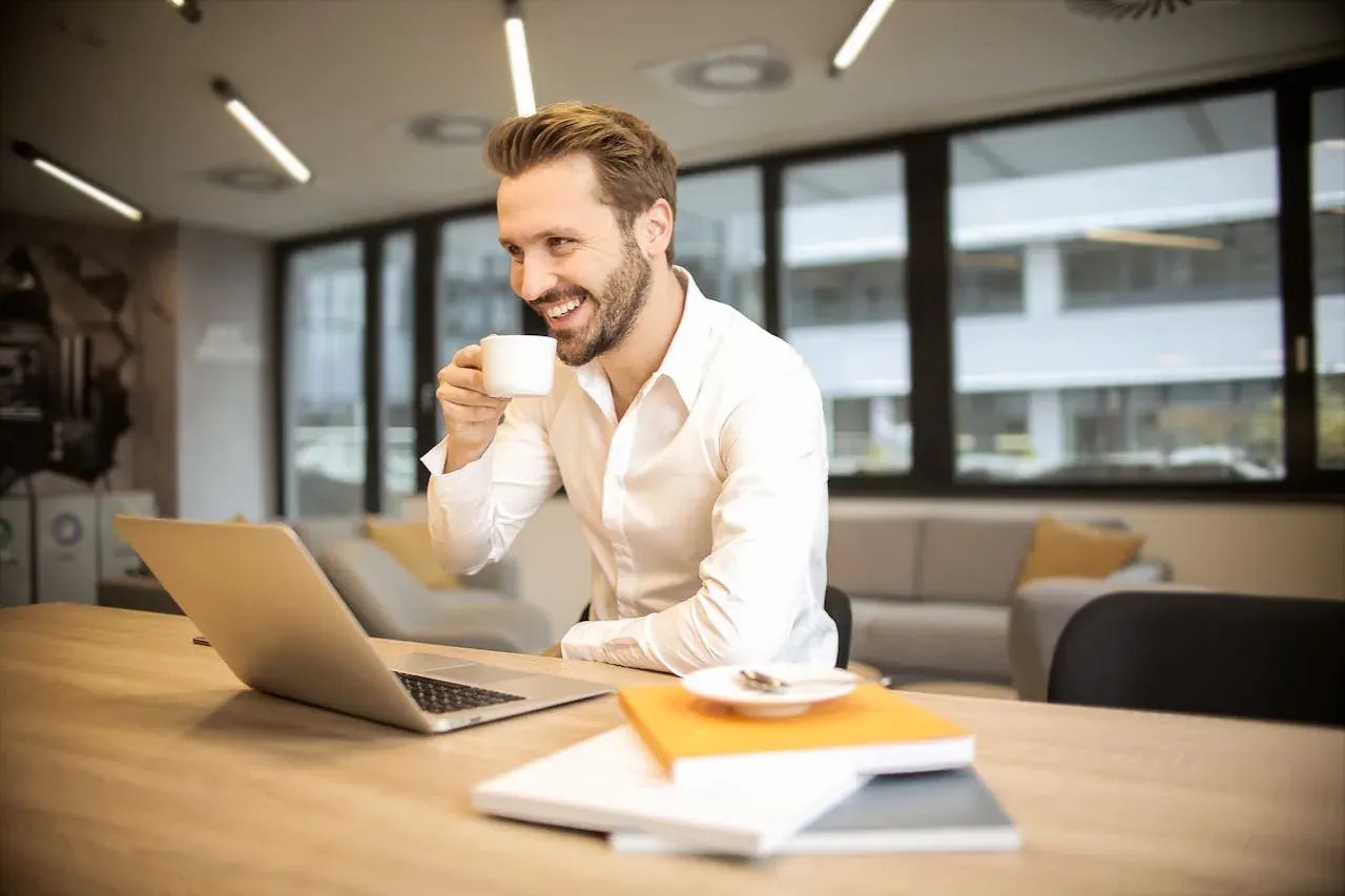 A smiling man in a white shirt sipping an espresso at a desk in a bright office lounge.