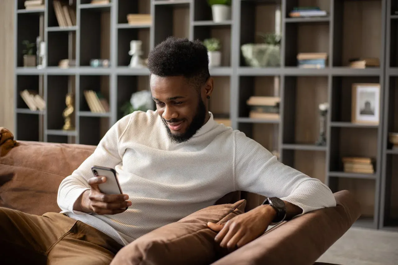 A bearded man in a cream sweater relaxing on a brown couch, smiling at his phone, with a full bookshelf behind him.