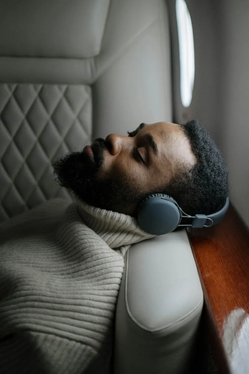 A bearded man resting with his eyes closed in a cream leather recliner, wearing headphones, a knit blanket pulled up to his chin.