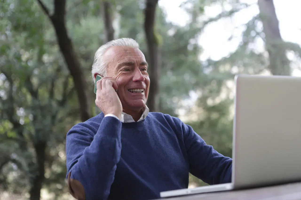 A gray-haired man in a navy sweater smiling as he talks on his phone, a laptop open in front of him.