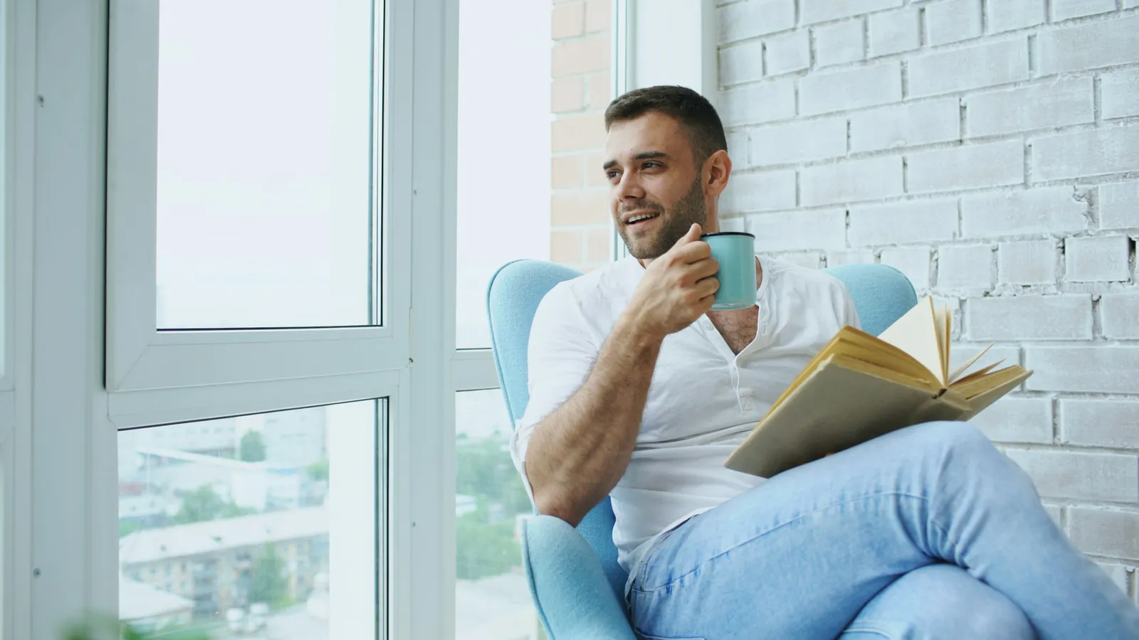 A man relaxing in a chair by a bright window with a book and a cup of coffee, smiling.