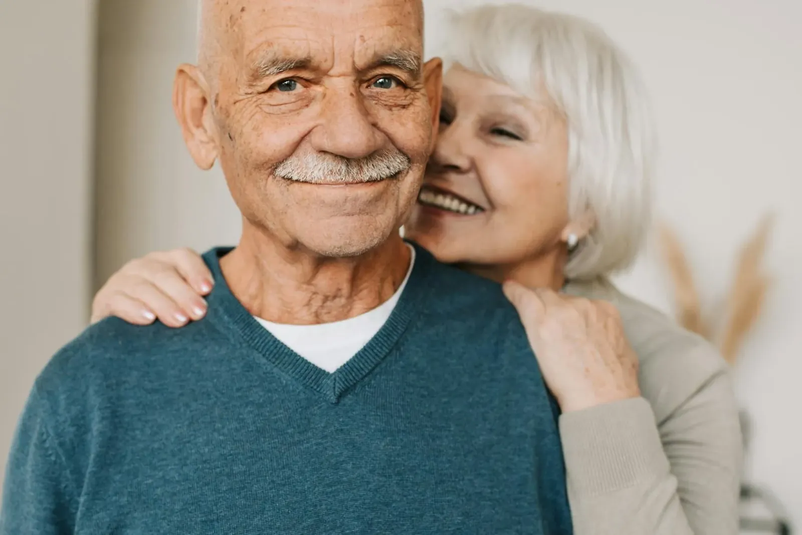 An older man smiling at the camera while his wife rests her cheek against his shoulder.