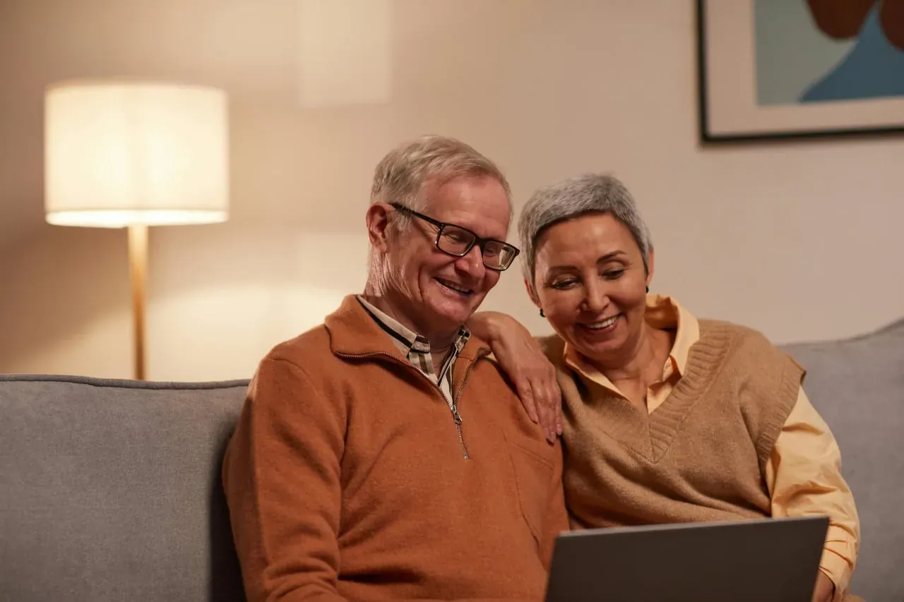 An older man and woman on a gray sofa smiling at a laptop, his arm around her shoulders.