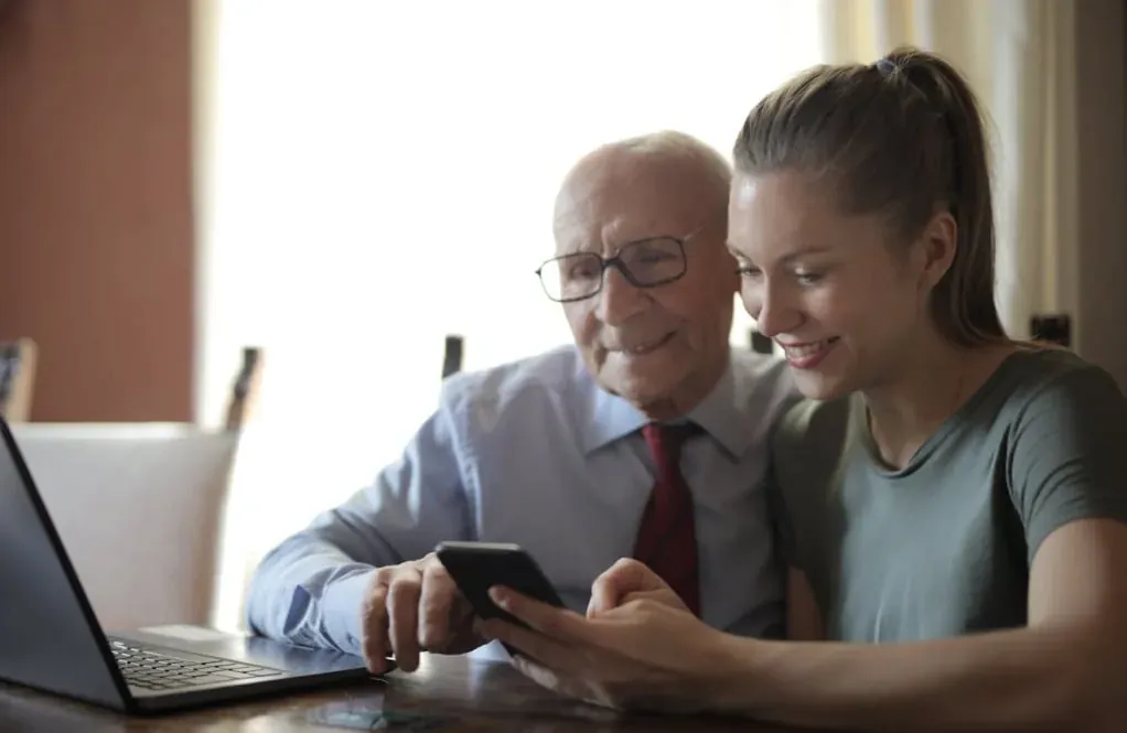An older man in glasses and a younger woman on a sofa, looking at and pointing to a screen together.
