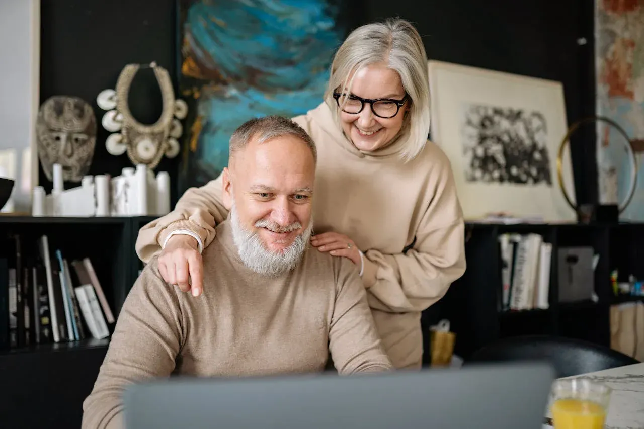 An older bearded man smiling at a laptop while an older woman stands behind him with a hand on his shoulder.