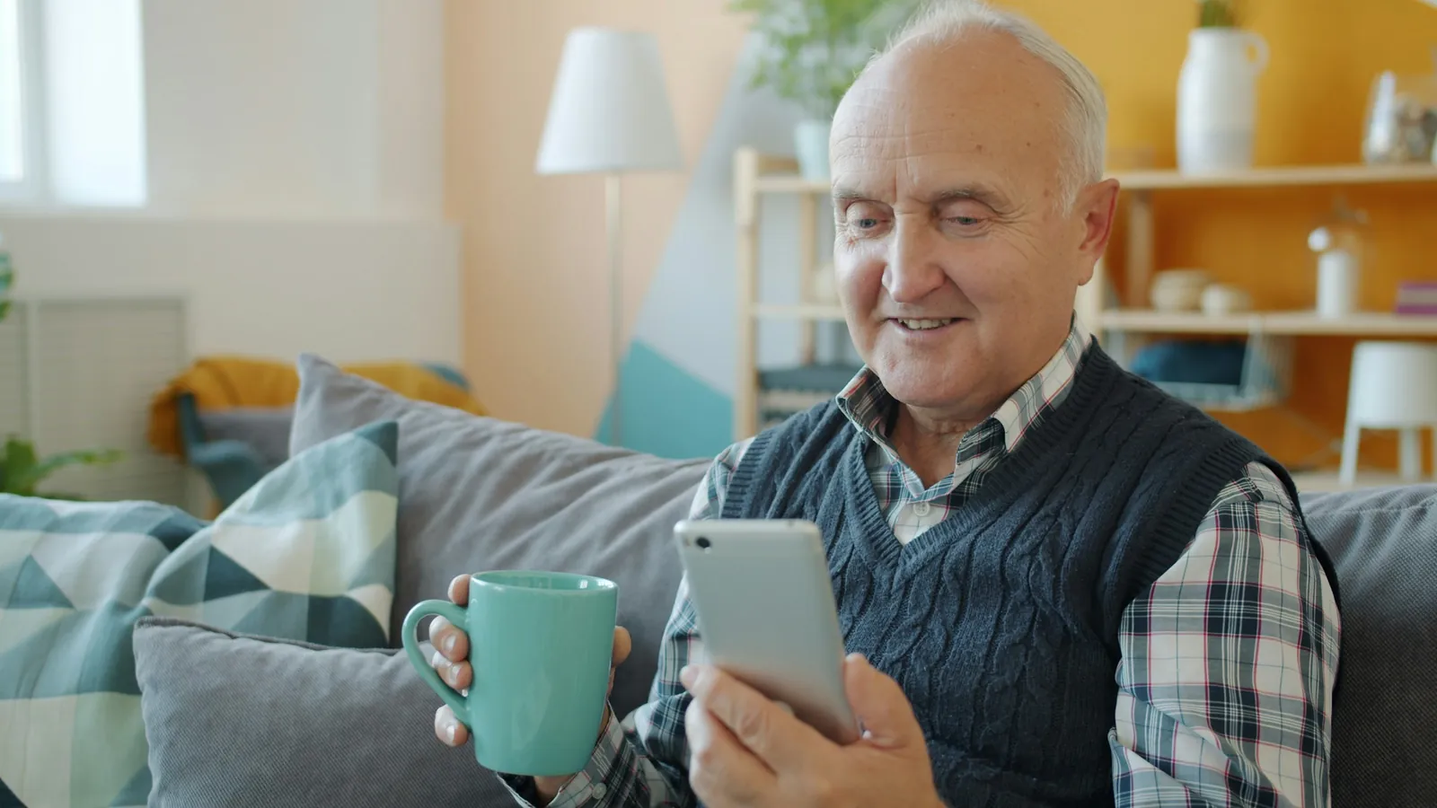 An older man on a sofa smiling at his phone, a mug in his other hand.