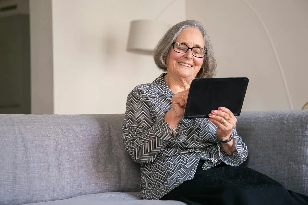 An older woman with curly gray hair and glasses smiling at a tablet on a gray sofa.