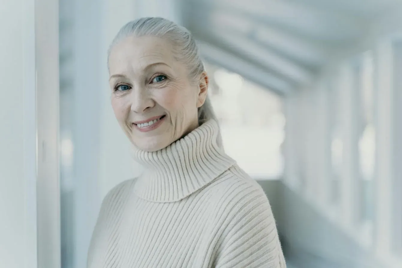An older woman with gray hair in a cream turtleneck smiling warmly in a bright hallway.