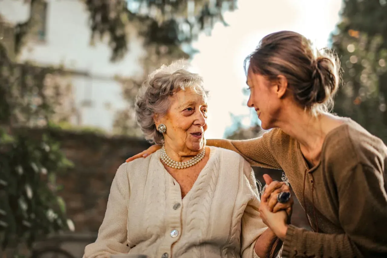 An older woman in a cream cardigan holding hands with a younger woman, both smiling warmly outdoors.