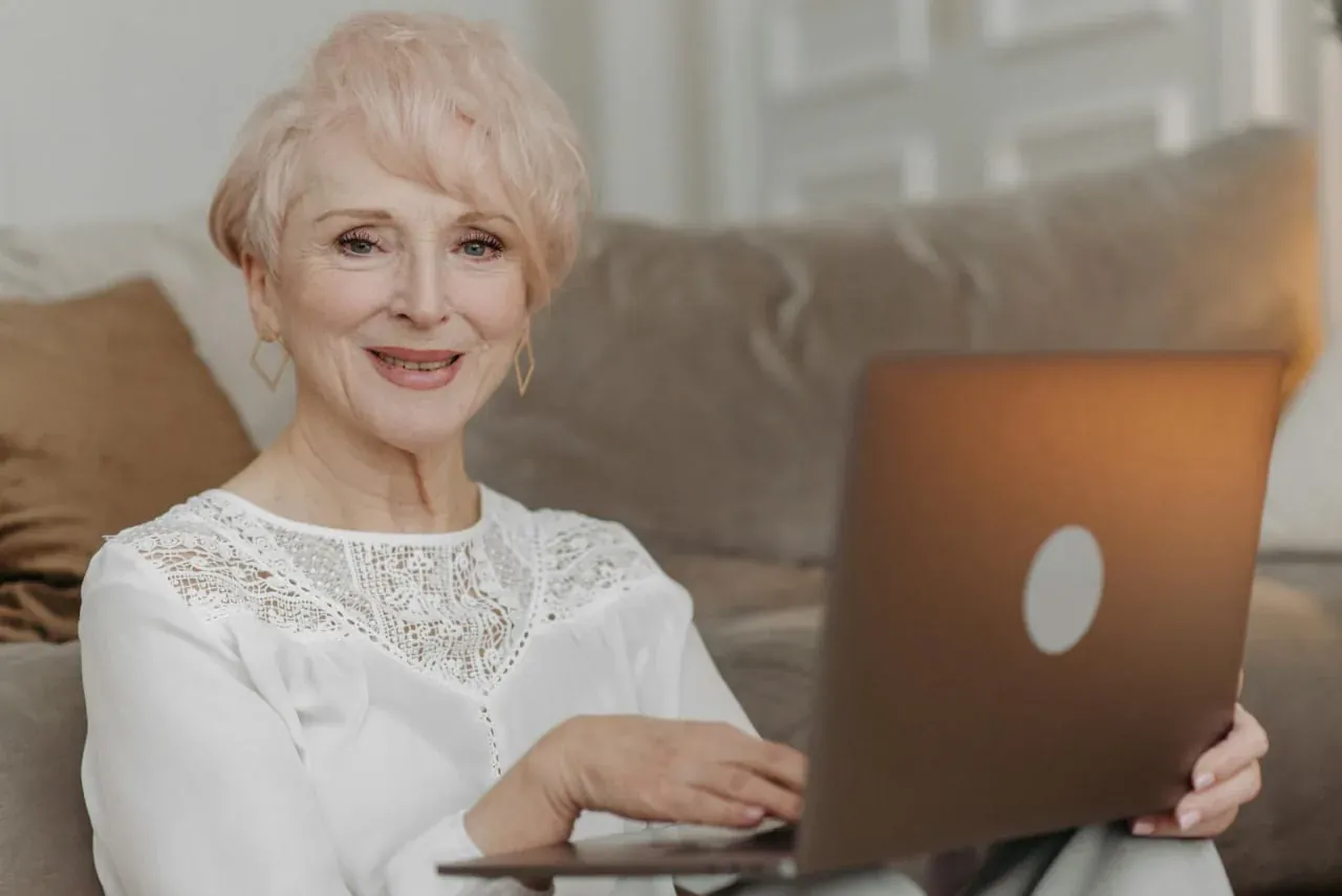An older woman with short blonde hair on a sofa, holding an open laptop and smiling.