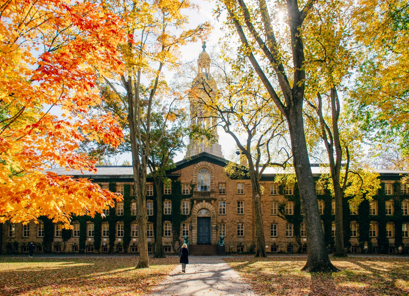 A person walking along a path toward a historic stone building framed by autumn trees.