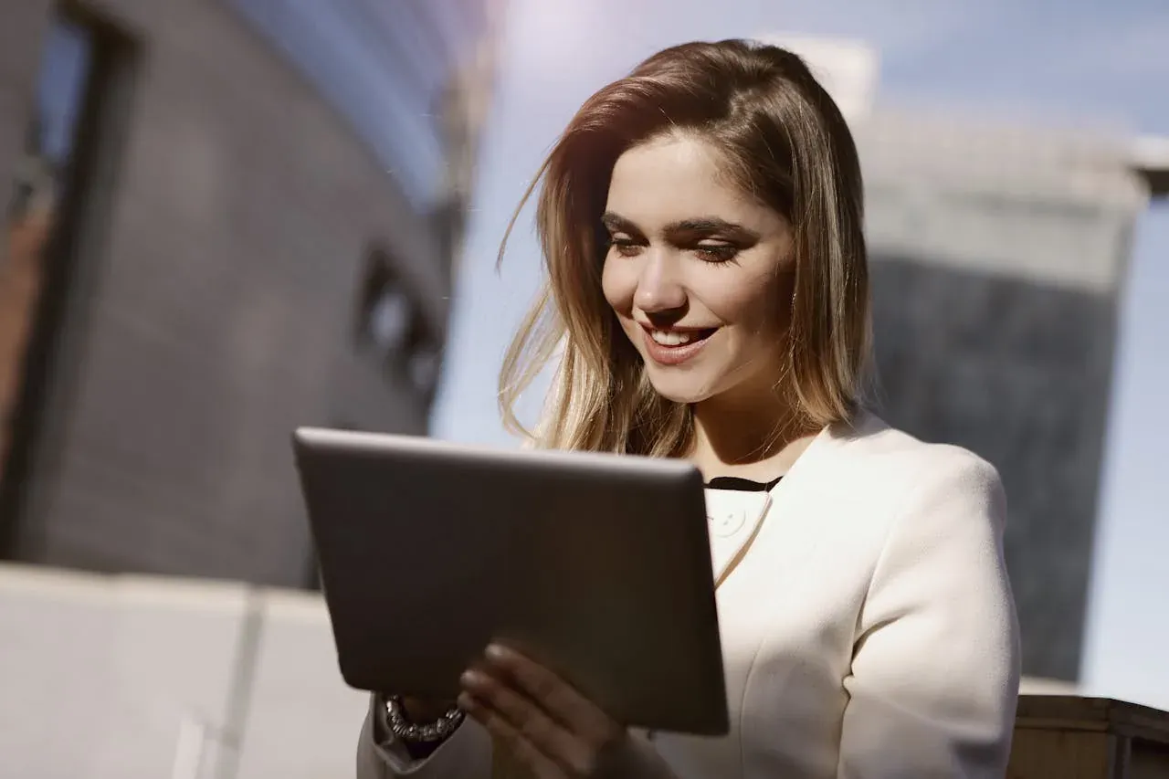 A young professional woman in a white blazer smiling down at a tablet outside modern glass office buildings.
