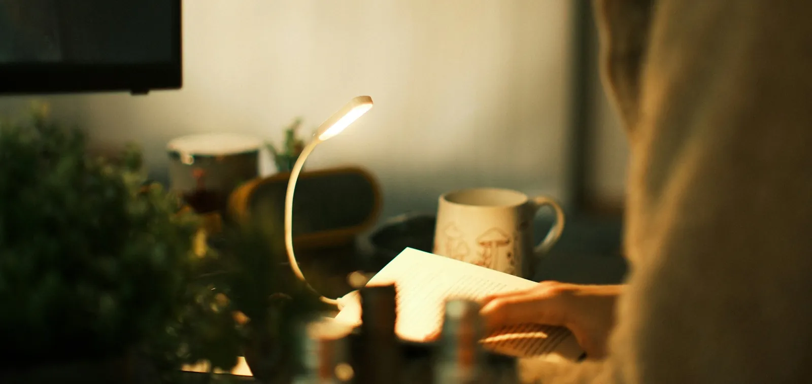 Someone reading at a desk by the light of a small lamp, a mug beside them.