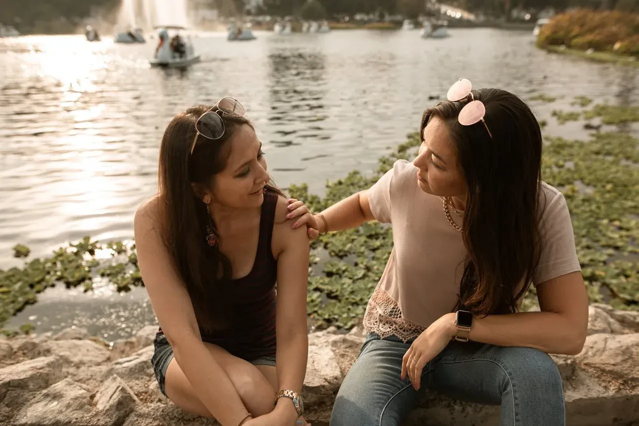 Two women sitting by a lake at sunset, one resting a comforting hand on the other's shoulder.