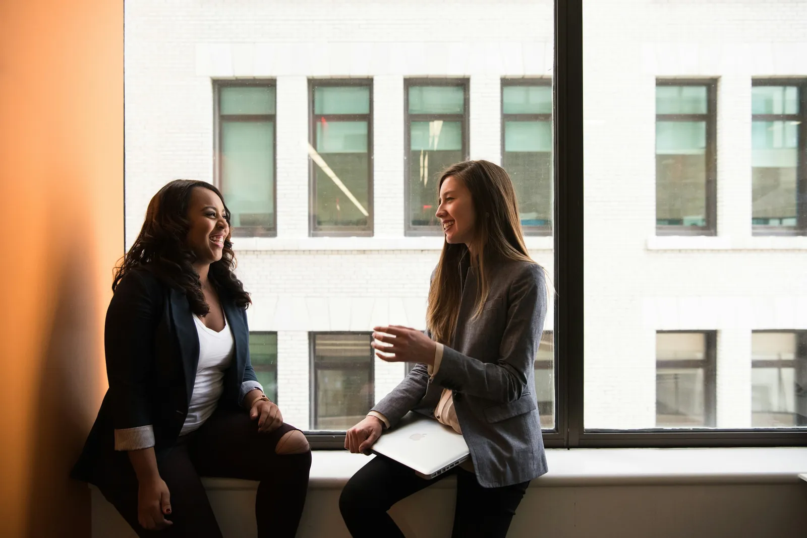 Two women sitting by a large window, laughing as they talk.