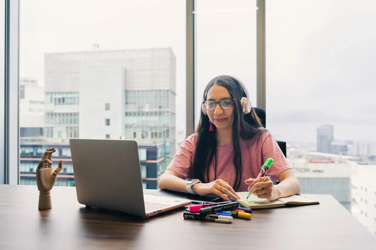 A woman in glasses and headphones working at a desk with notes and markers, a city skyline behind her.