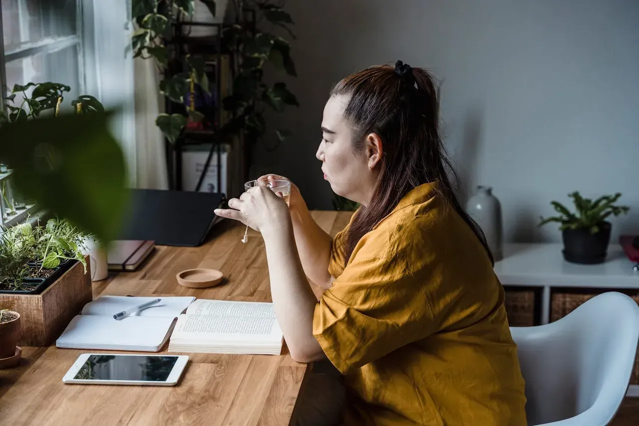 A woman in a mustard linen shirt sipping from a small glass at a wooden desk surrounded by plants.