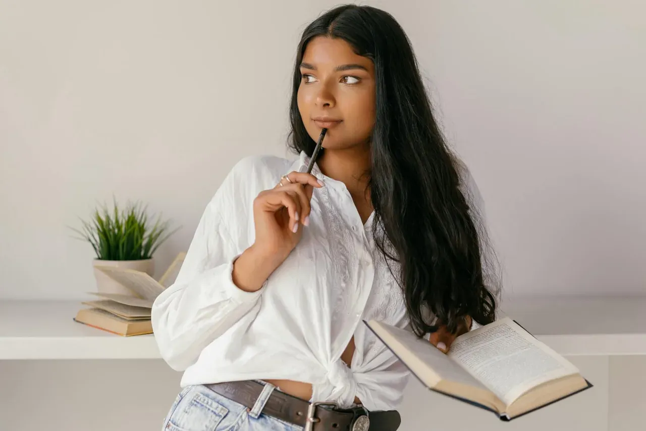 A young woman in a white blouse holding a pen to her chin and an open book, thinking.