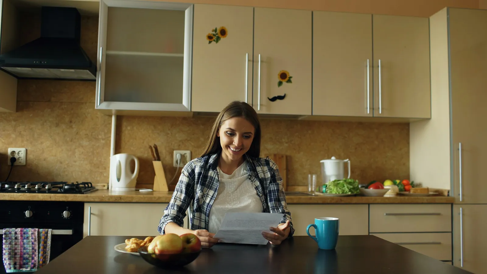 A woman at her kitchen table smiling as she reads a letter, a mug of coffee beside her.