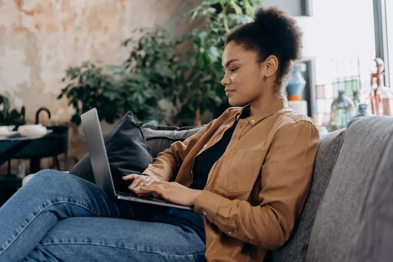 A woman with curly hair sits cross-legged on a gray couch in a bright, plant-filled apartment, typing on a laptop on her knees.