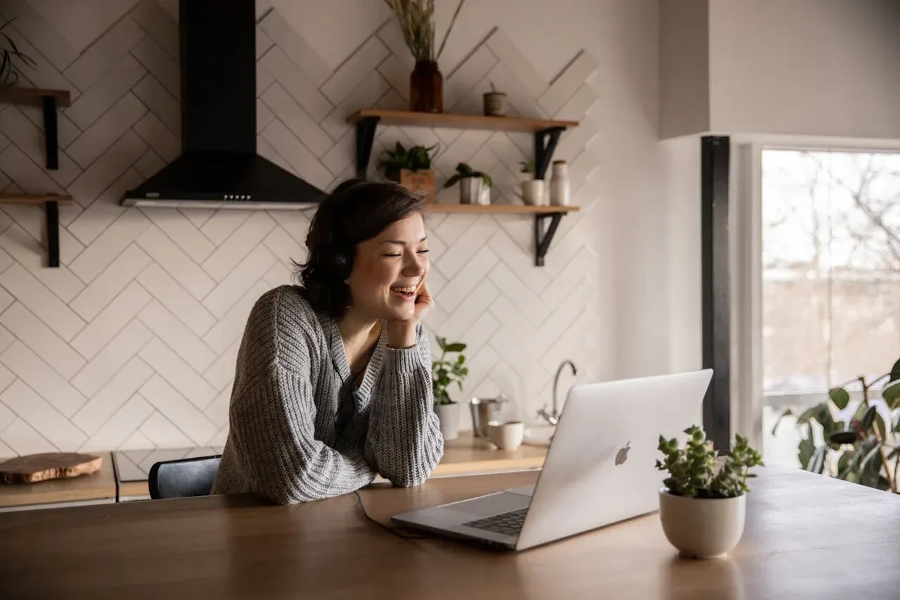 A woman in a gray sweater, wearing headphones, laughs as she looks at her laptop at a bright kitchen table.