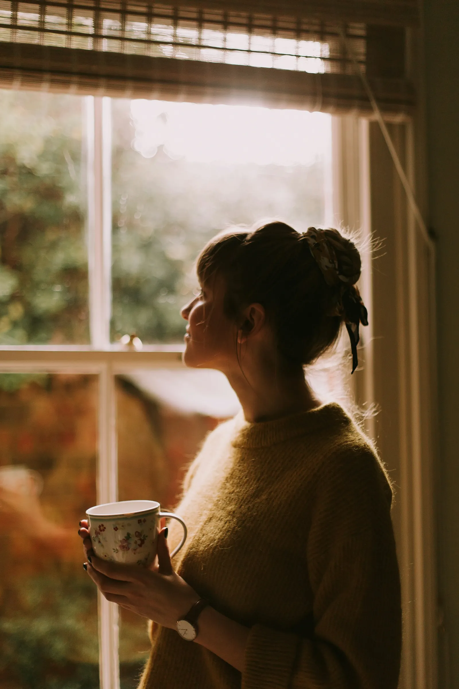 A woman holding a mug, looking out of a sunlit window.