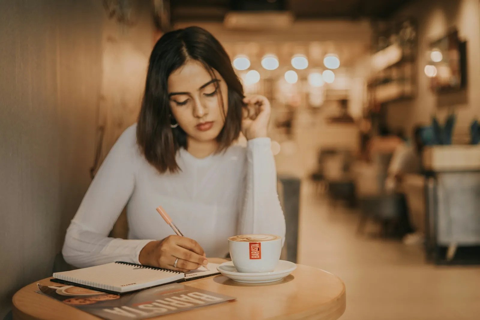 A woman writing in a notebook at a small café table, a cup of coffee in front of her.