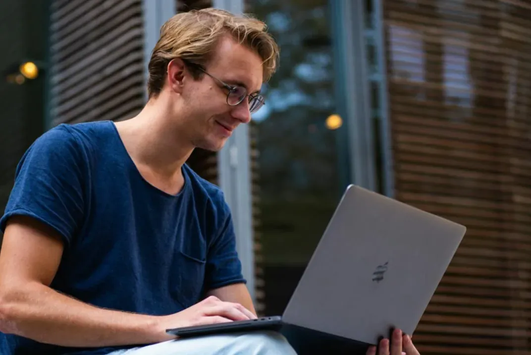 A young man with glasses smiling at a laptop on his lap, sitting outdoors.